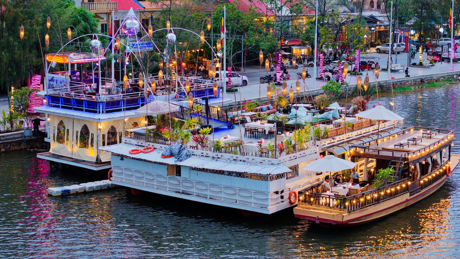 Wide dusk view of the Kdam Kampot boats and floating bar glowing along the Kampot river