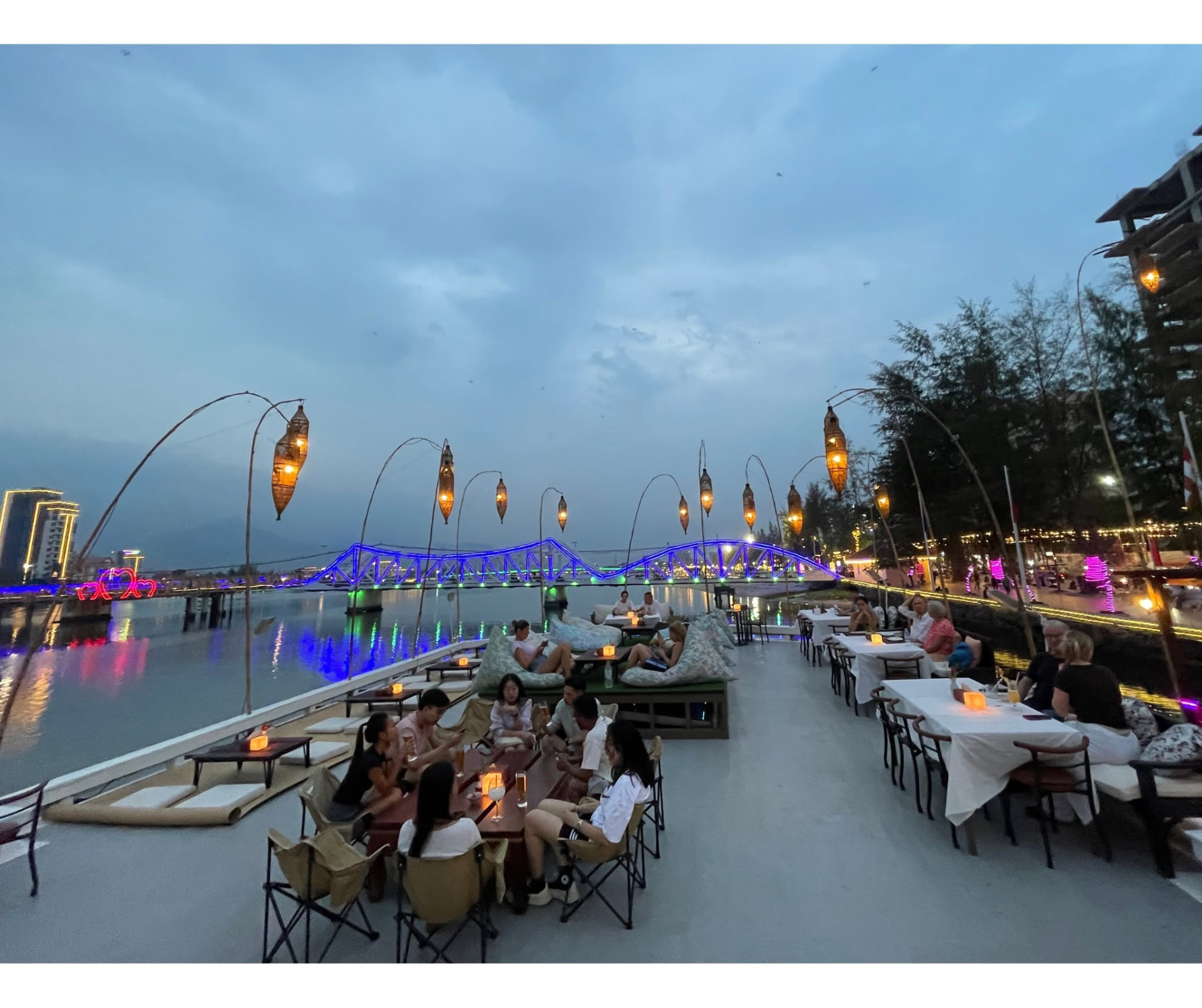 Guests dining on the Kdam Kampot riverside deck at dusk with the lit Old Bridge behind