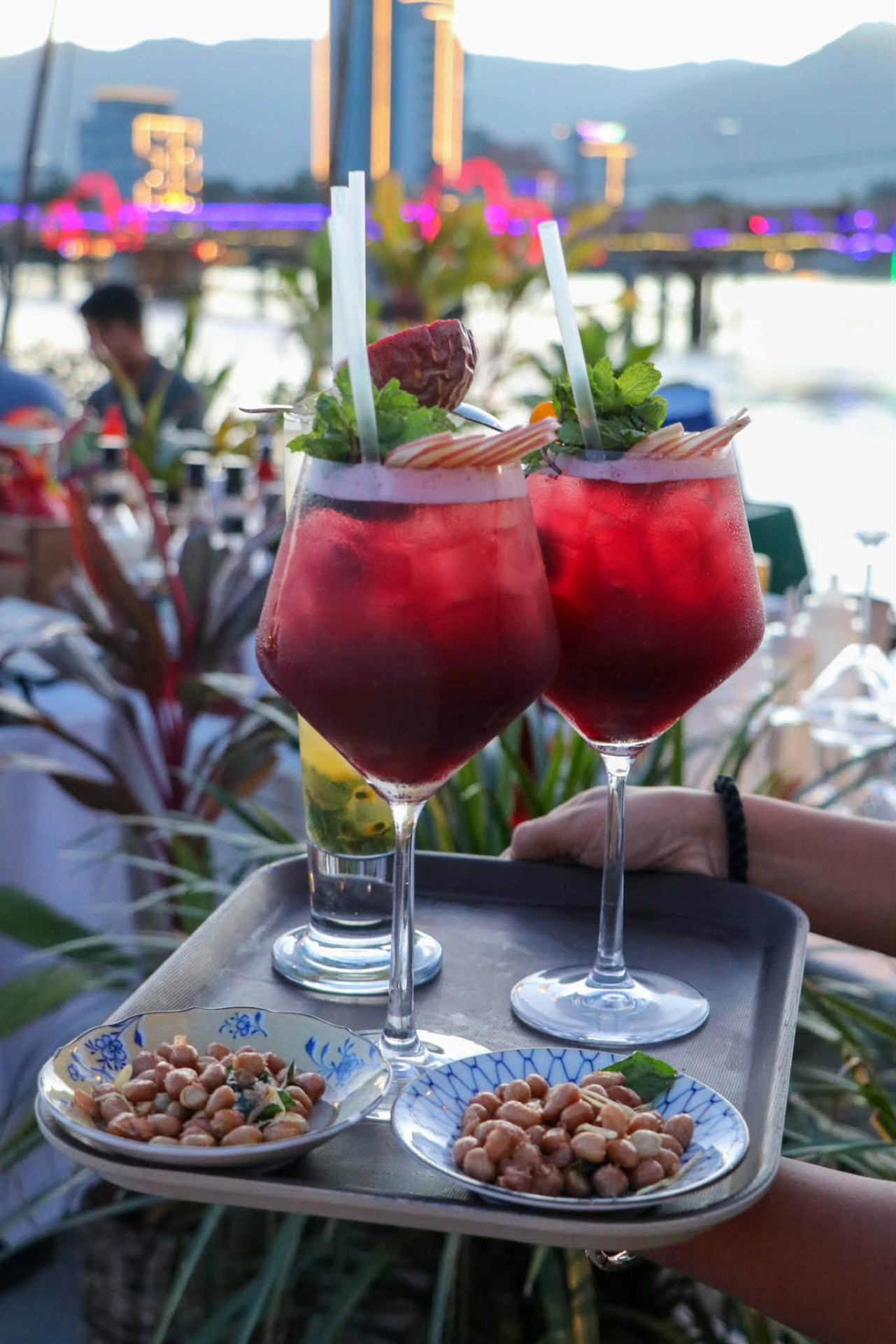 Ruby cocktails and roasted peanuts on a tray with the lit riverside behind