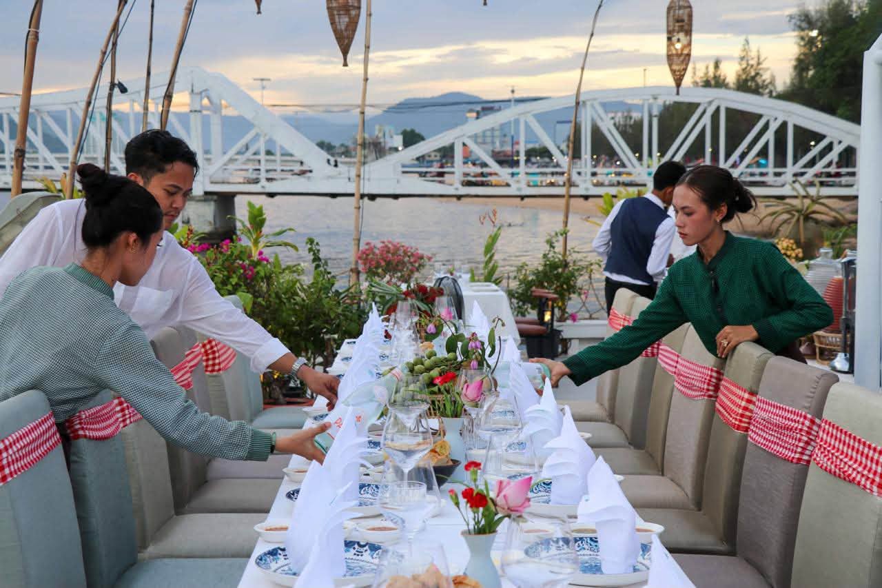 Kdam Kampot team setting a long banquet table facing the white Old Bridge at dusk